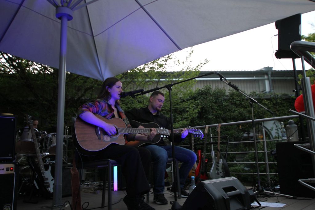 Zwei Musiker mit Akustikgitarren spielen auf einer Bühne unter einem Sonnenschirm.