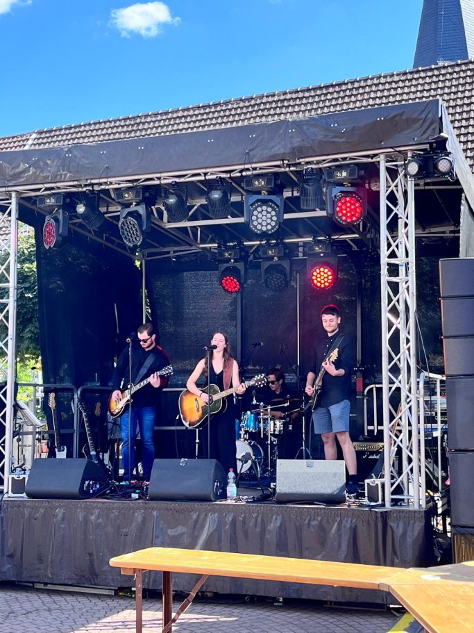 Eine Band spielt auf einer Bühne, umgeben von Lichtern und einem Publikum im Freien.